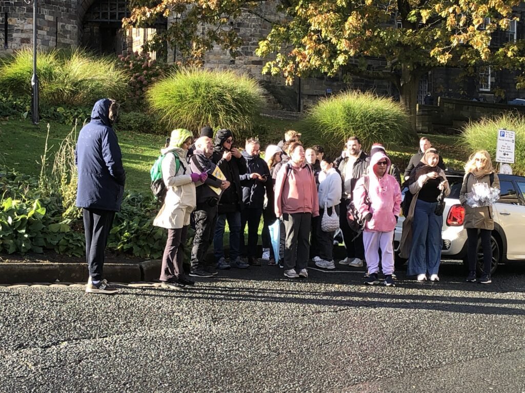 A group of people, on a guided walks in Lancaster, some wearing jackets and hoodies, stand on a pavement by a road in bright sunlight near Castle Hill, with greenery and Lancaster Castle in the background.